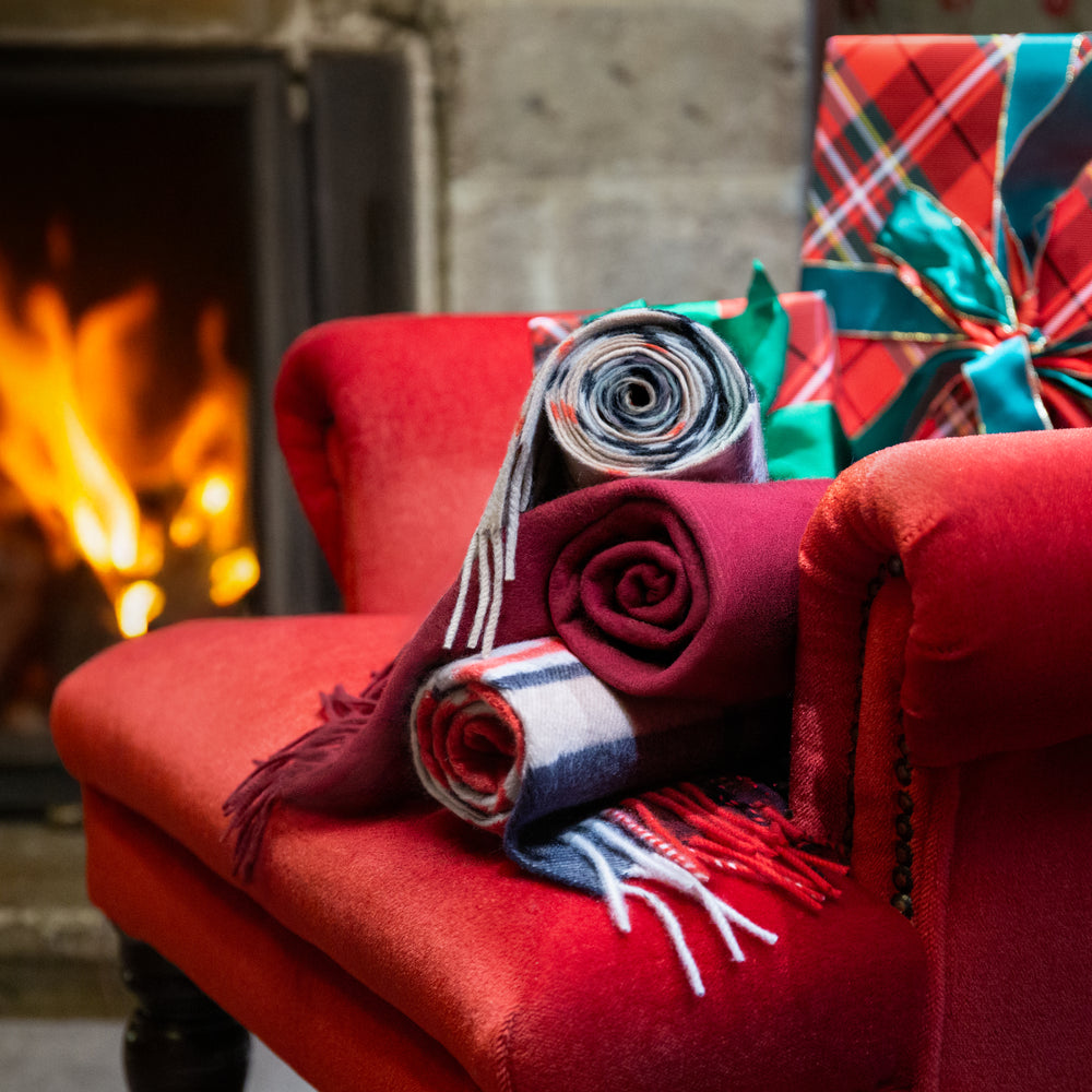 A cosy holiday scene with rolled plaid and plain cashmere scarves draped over a red armchair beside a fireplace, with wrapped Christmas gifts in the background.