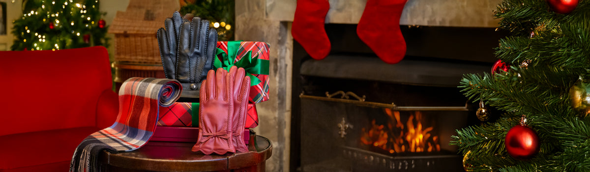 A cosy holiday scene with a round table displaying a pair of men's black leather gloves, a pair of women's red leather gloves, a cashmere plaid scarf, and a wrapped gift, set in front of a fireplace with red stockings and a Christmas tree.