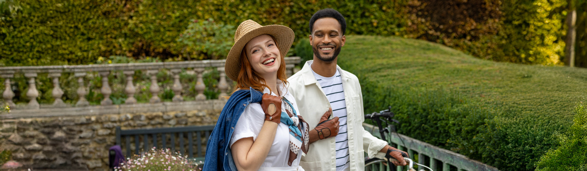 A smiling couple posing outdoors in a garden setting. Wearing leather gloves in brown shades. 